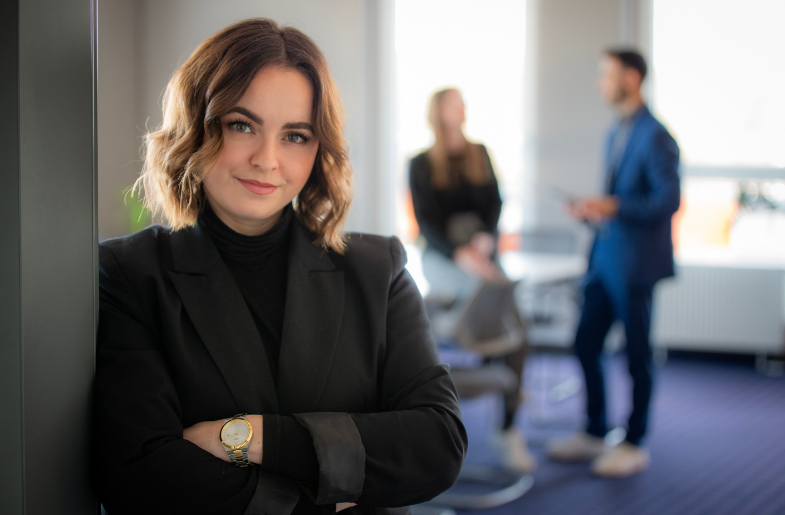 Fokus auf eine Person im Vordergrund, die sich mit verschränkten Armen an einen Türrahmen lehnt, im Hintergrund sind verschwommen zwei Personen im Gespräch zu erkennen.